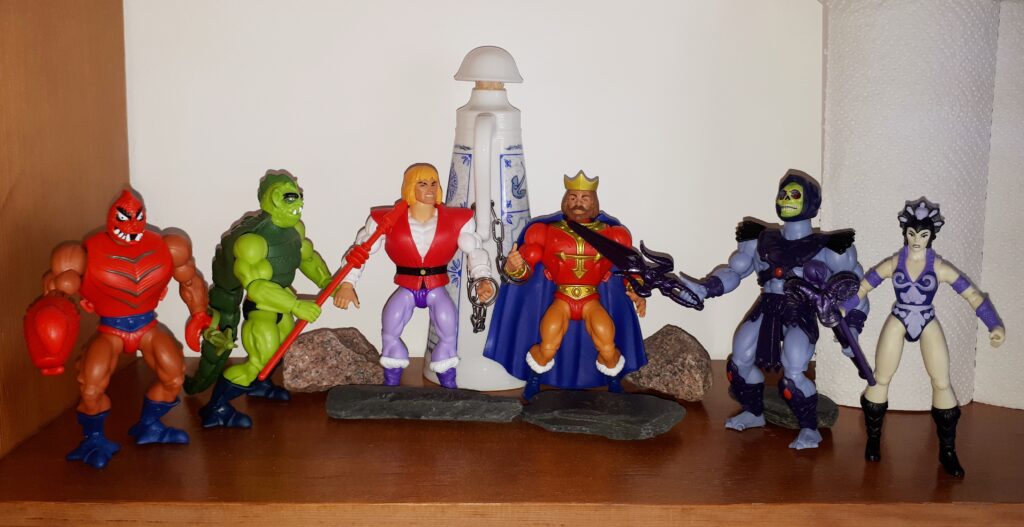 Prince Adam looks questioningly at his father, while Skeletor and his Evil Warriors threaten them both