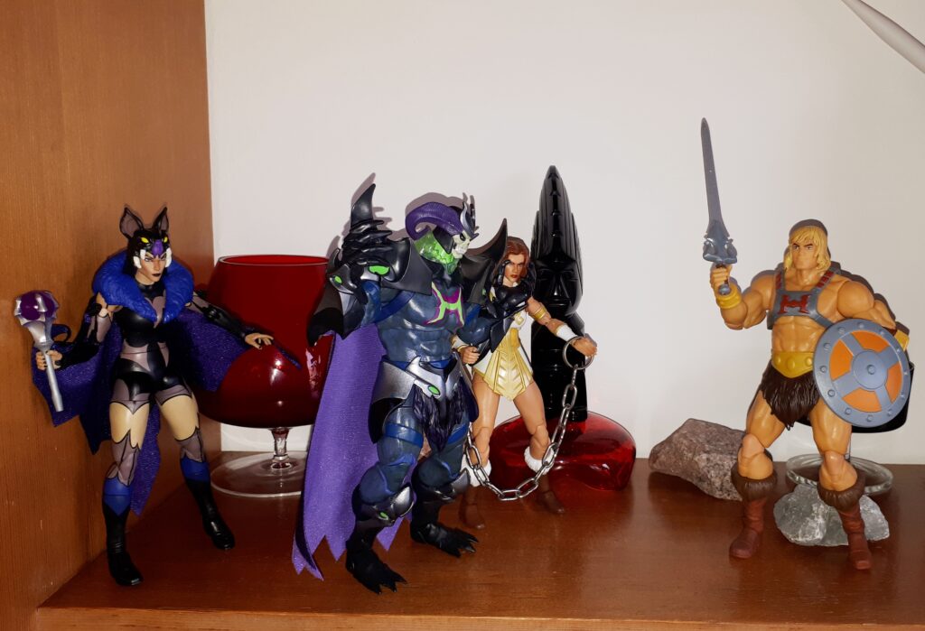 Skelegod threatens the chained up Teela in front of He-Man, while Evil-Lyn looks on.