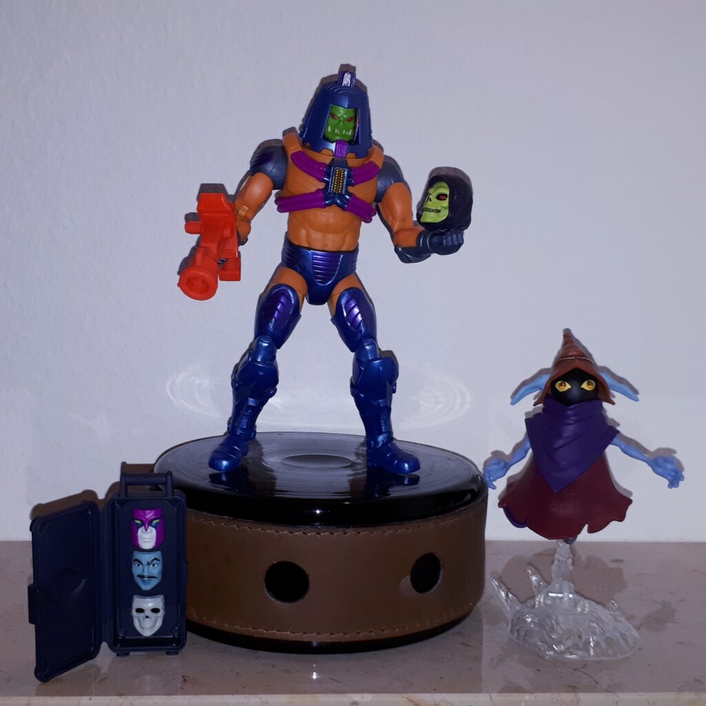 Man-e-Faces is wearing his monster face and holds Skeletor's skull, while Orko looks on.