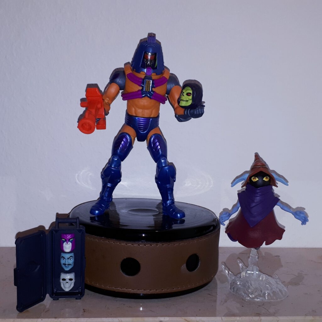 Man-e-Faces wears his robot face and holds Skeletor's skull, while Orko looks on.